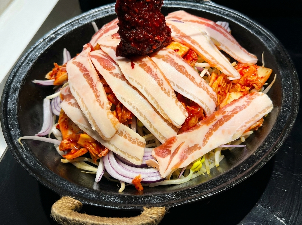Eine leuchtend rote koreanische Pfannensauce wird über eine Pfanne mit geschichtetem Schweinebauch und frischem Gemüse verteilt.
