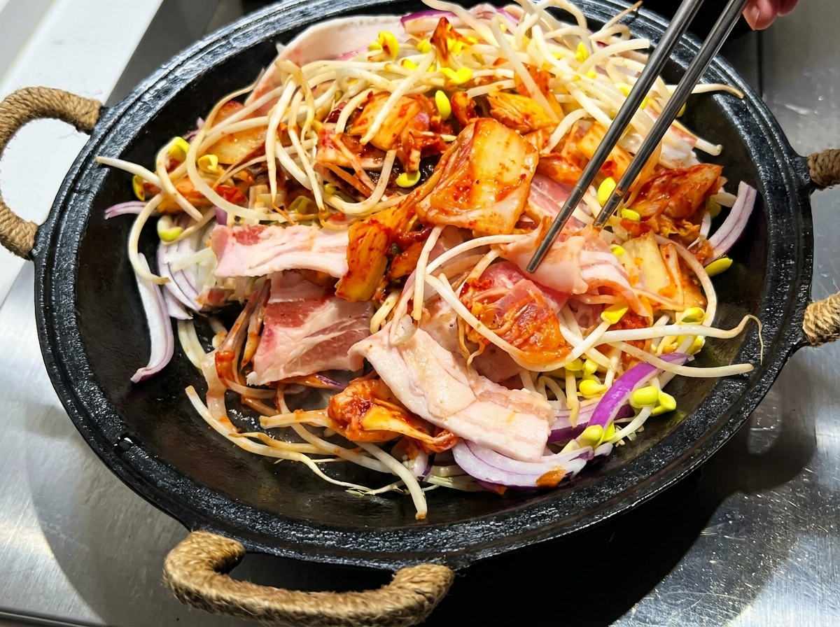Eine Zange vermischt Schweinebauch, Sojasprossen, Kimchi und Sauce in einem gusseisernen Topf.