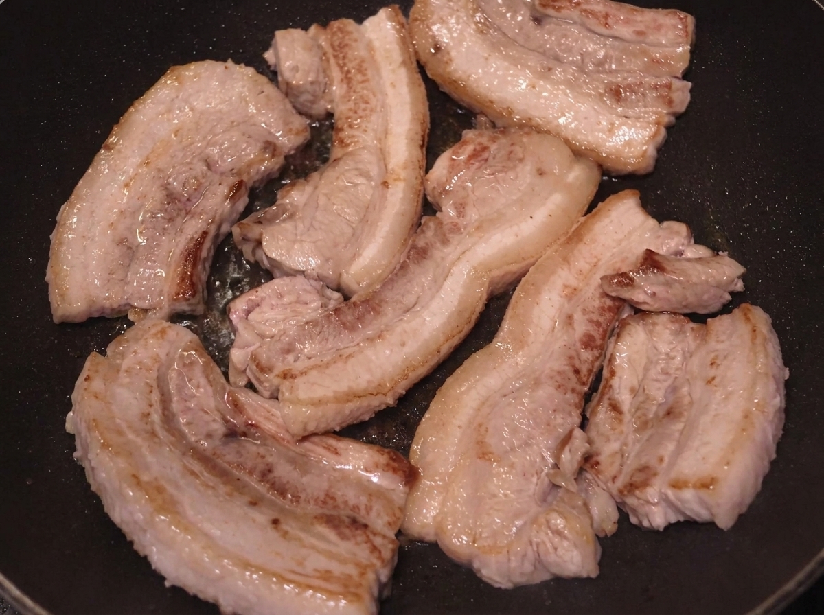 Mehrere Schweinebauchscheiben brutzeln in einer Pfanne, die Oberflächen sind tief gebräunt.