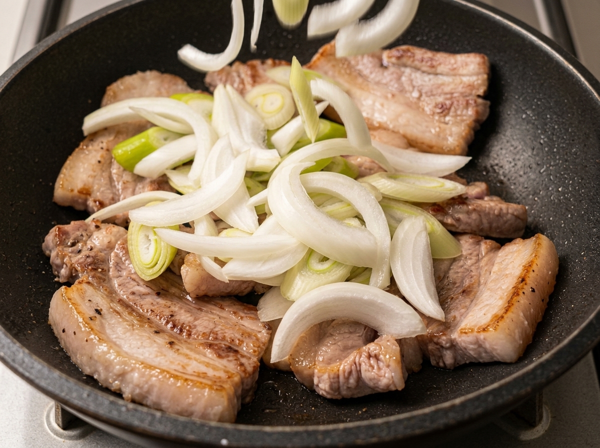 In Scheiben geschnittene weiße Zwiebeln und Frühlingszwiebeln, verstreut über gebräunten Schweinebauchscheiben in einer Pfanne.