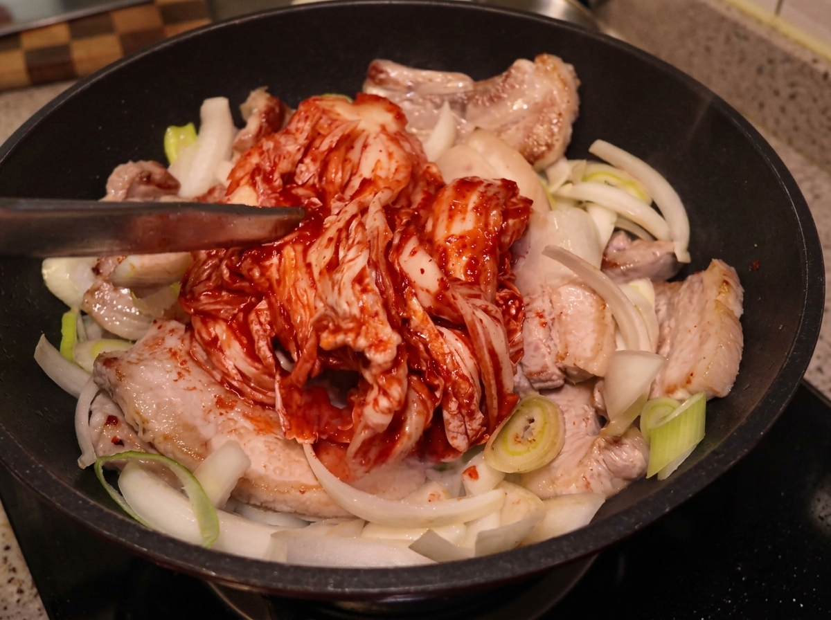 Ein großes, ganzes Stück saures Kimchi wird mit einer Zange auf den angebratenen Schweinebauch und die Zwiebeln in eine Pfanne gelegt.