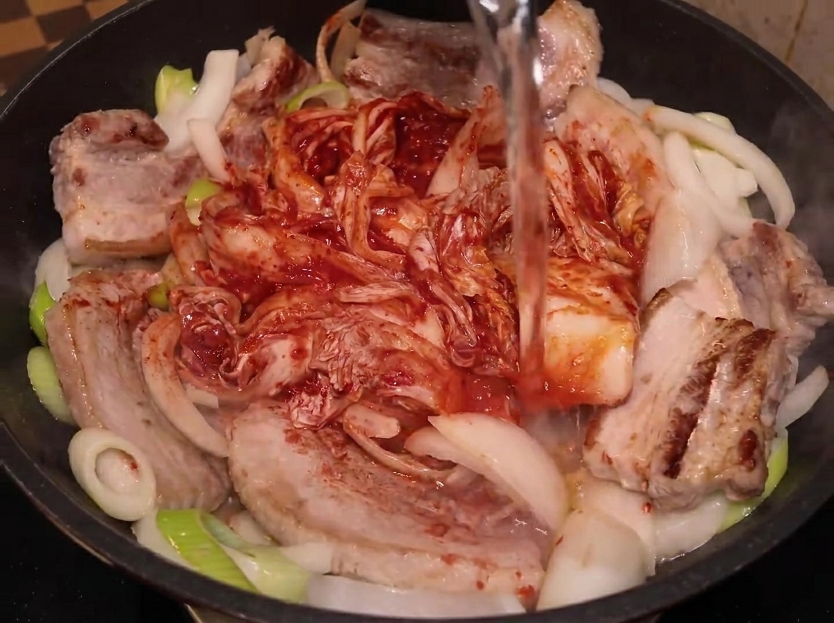 Wasser wird aus einer Plastikflasche in eine Pfanne mit Schweinebauch, Zwiebeln und ganzem Kimchi gegossen.