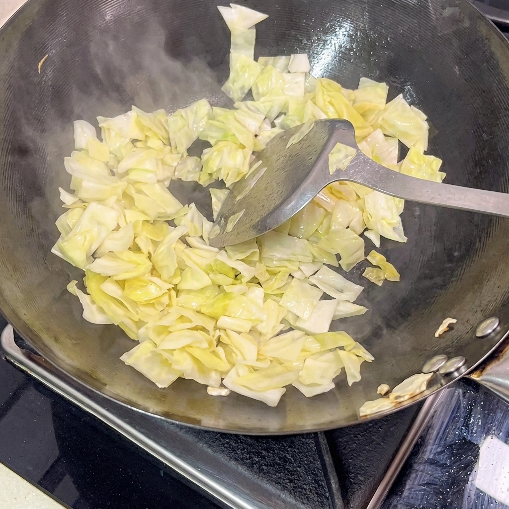 Ein Metallspatel wendet hellgrünen eingelegten Kohl in einem heißen, schwarzen Wok, sichtbarer Dampf steigt auf.