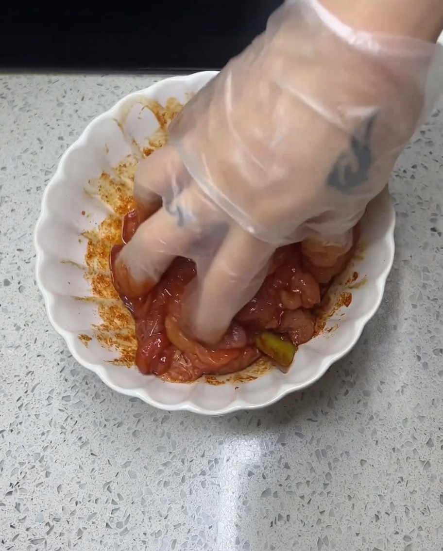 Eine behandschuhte Hand massiert eine rötliche Marinade in rohe Hähnchenstücke.
