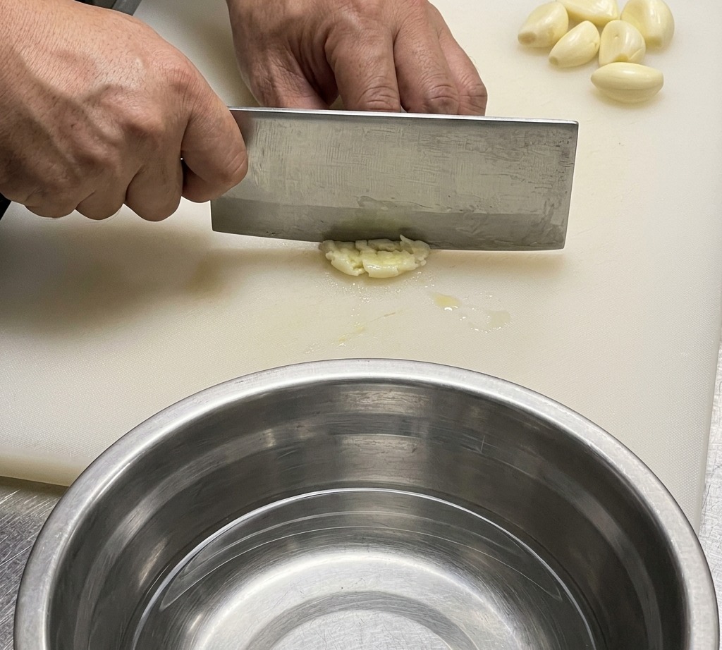 Ein Kochmesser schneidet Knoblauch und Ingwer auf einem weißen Schneidebrett.