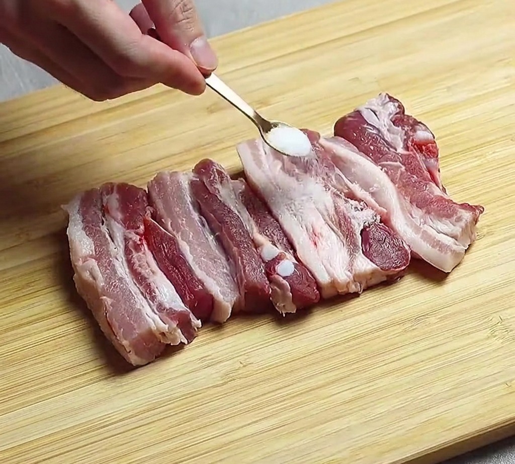 Auf einem Holzbrett ausgelegte rohe Schweinebauchstreifen werden mit einer Prise Salz bestreut.
