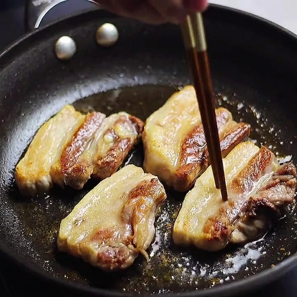 Goldbraune Schweinebauchstreifen braten in der Pfanne; mit einem Stäbchen wird geprüft, ob das Fleisch gar ist.
