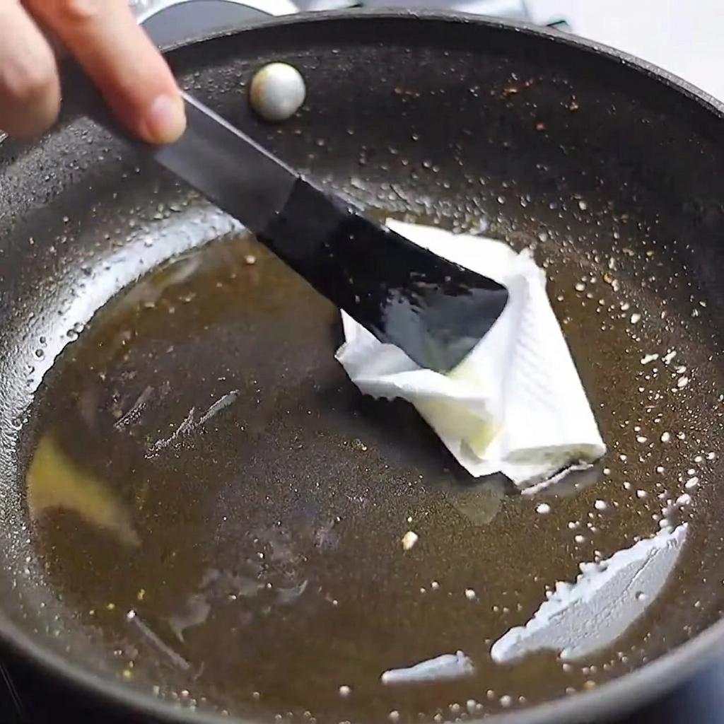 Ein mit einer Zange gehaltenes Küchentuch wird verwendet, um überschüssiges Öl aus einer Pfanne zu wischen.
