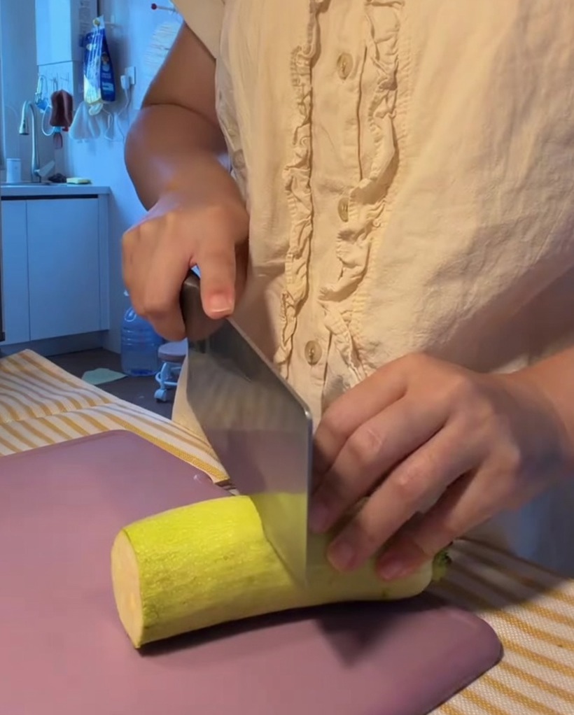 Frische Zucchini wird auf einem lilafarbenen Schneidebrett in gleichmäßige Halbmonde geschnitten.