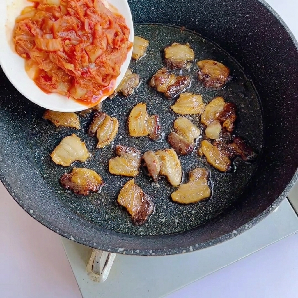 Gehacktes Kimchi wird zu den angebratenen Schweinebauchscheiben in die Pfanne gegeben.