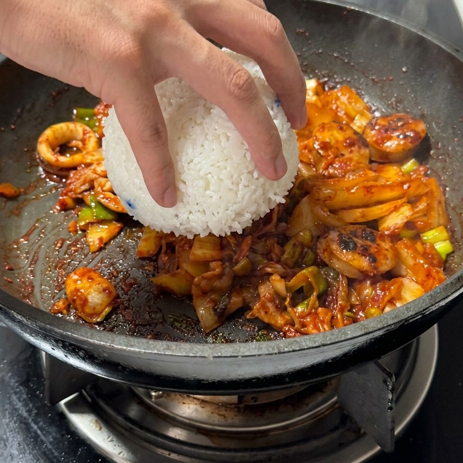 Eine Schüssel mit weißem, gedämpftem Reis wird zu der angebratenen Kimchi-Fleisch-Mischung in die Pfanne gegeben.