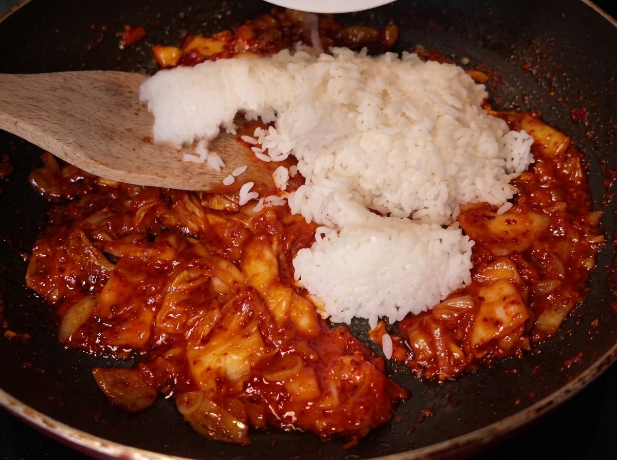 Ein Holzspatel gibt eine große Portion weißen Reis in die Pfanne, in der bereits die rote Kimchi-Mischung brät.