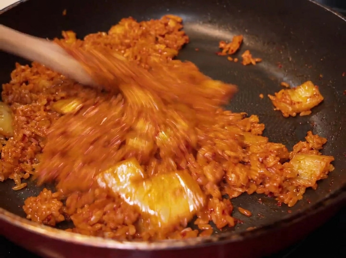 Eine Nahaufnahme des Reises, der in der Pfanne mit dem roten Kimchi vermengt wird und eine gleichmäßige rötlich-orange Farbe annimmt.