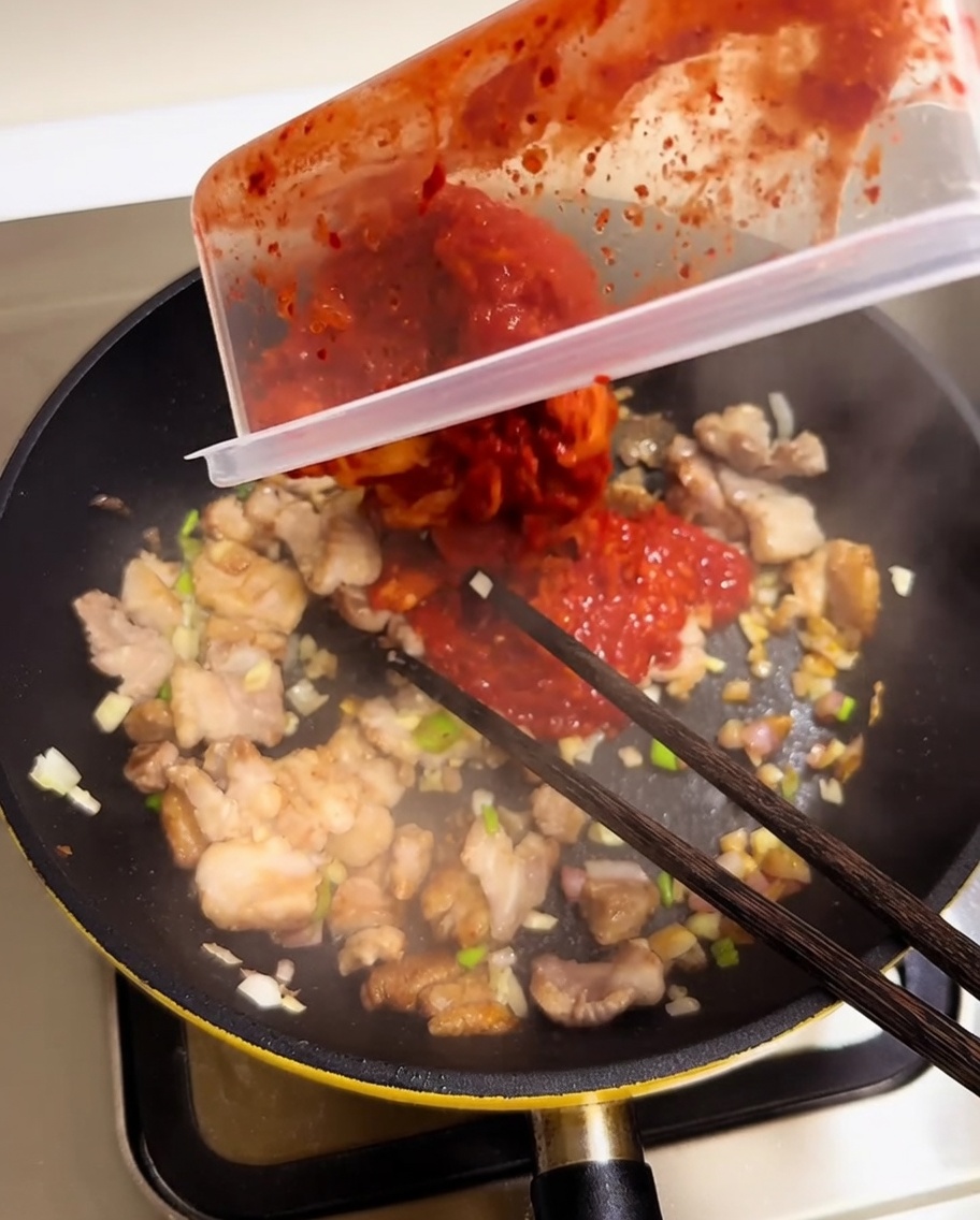 Gehacktes rotes Kimchi wird aus einem Behälter in eine Pfanne mit Fleisch und Aromaten gegeben.