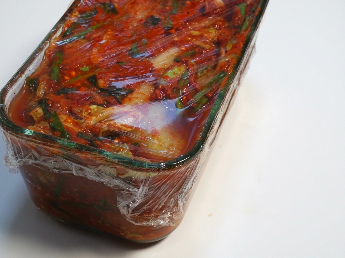 Behälter mit fermentiertem Kimchi, bei dem die Frischhaltefolie zurückgezogen wird.