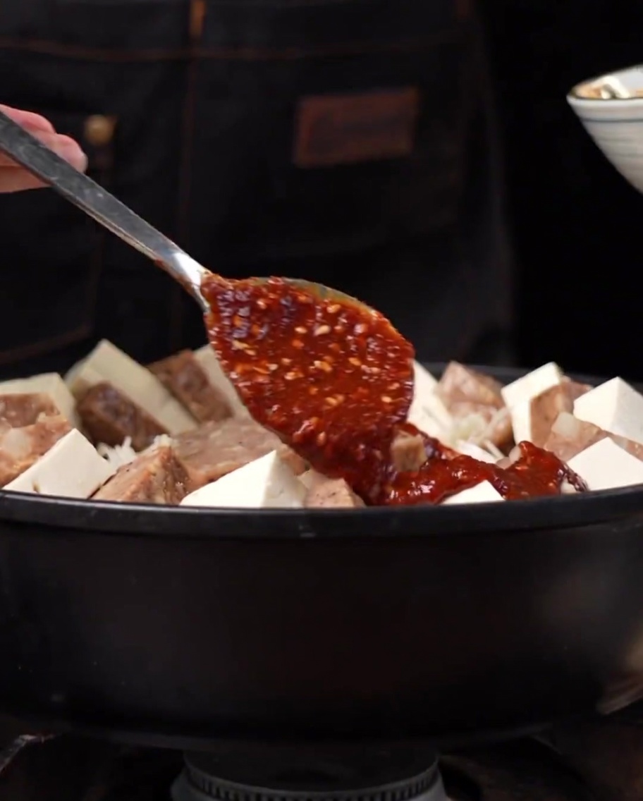 Eine dicke rötliche scharfe Sauce wird über die arrangierten Zutaten in einem Metalltopf gelöffelt und verteilt.