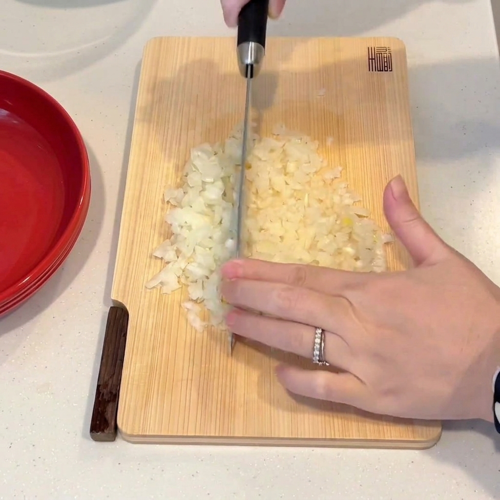 Eine Hand schneidet mit einem Kochmesser eine Zwiebel auf einem Holzbrett in feine Würfel.