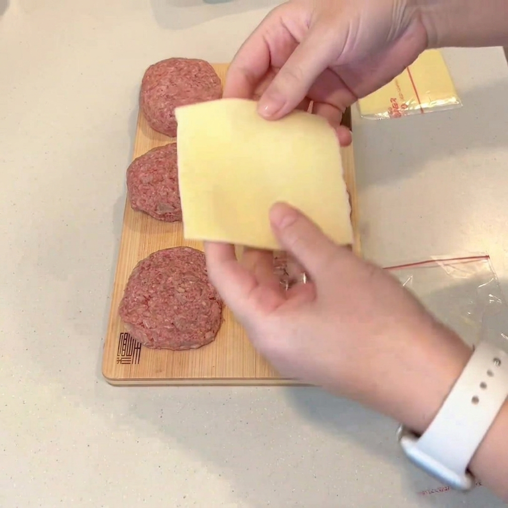 Eine gefaltete Käsescheibe wird in die Mitte eines flachgedrückten, rohen Hacksteaks gelegt.