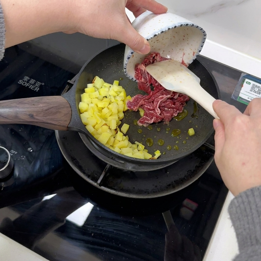 Rindfleischscheiben werden mit einem Spatel zu den gebratenen Kartoffeln in die Pfanne gegeben.