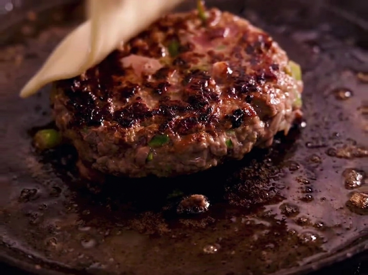 Eine Scheibe weißer Käse schmilzt auf einem gebratenen Burger-Patty in einer gusseisernen Pfanne.