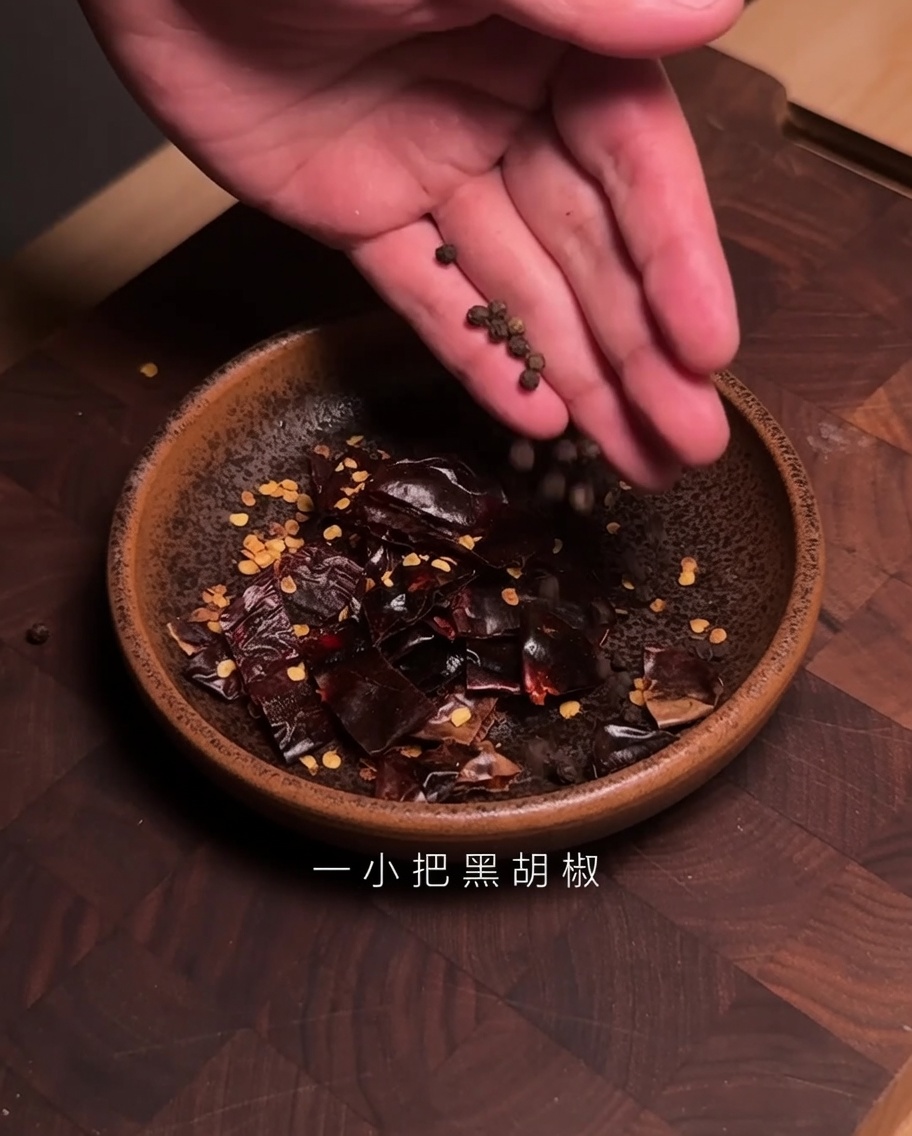 Hände geben schwarze Pfefferkörner in eine Schüssel mit zerkleinerten Guajillo-Chilis.