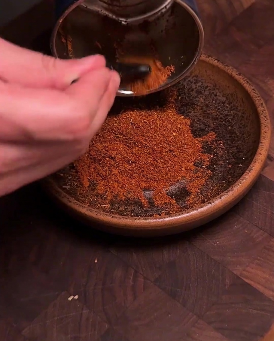 Eine Gewürzmühle gießt frisch gemahlenes, tiefrotes Chili- und Gewürzpulver in eine braune Keramikschüssel.