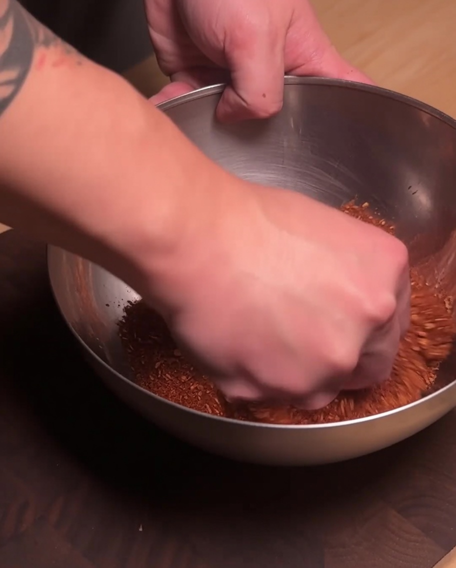 Eine Hand mischt das gewürzte Schweinehackfleisch und das rote Gewürzpulver in einer Edelstahlschüssel.