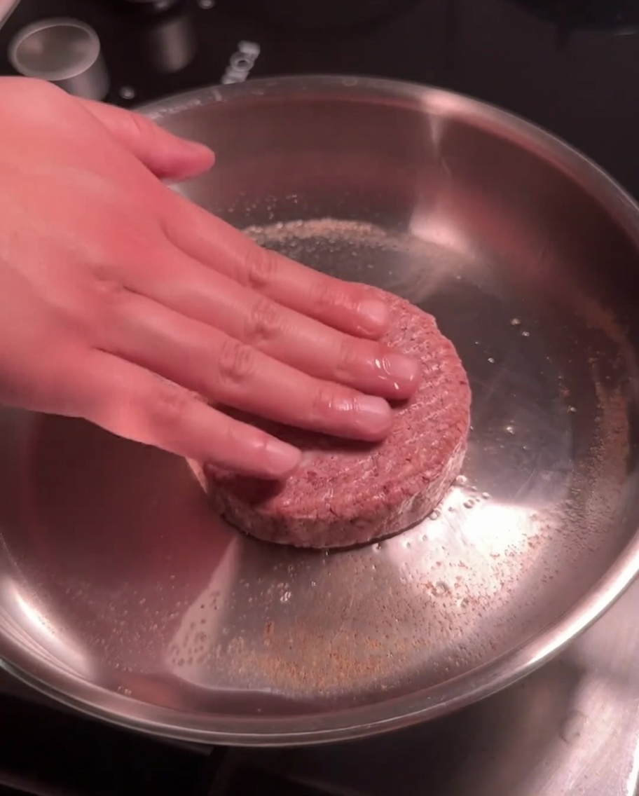 Ein rohes Rindfleisch-Patty wird zum Anbraten in eine heiße Edelstahlpfanne gedrückt.