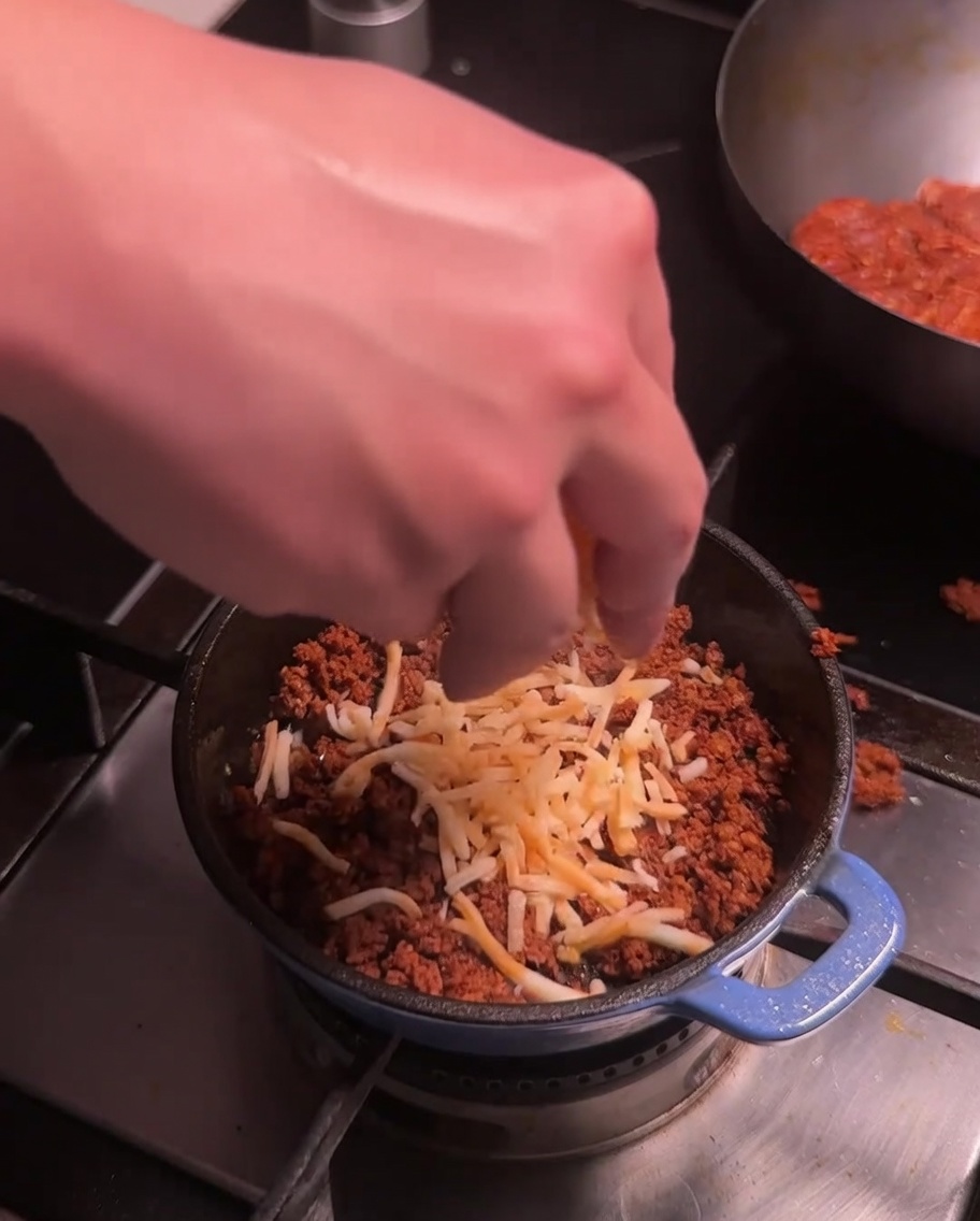Geriebener Käse wird über das gebratene Chorizo-Fleisch in einem kleinen blauen Topf gestreut.
