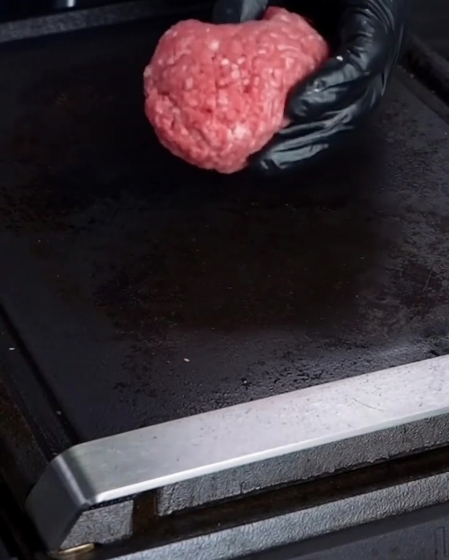 Eine behandschuhte Hand legt eine Kugel rohes 75/25 Rinderhackfleisch auf eine schwarze Grillplatte.