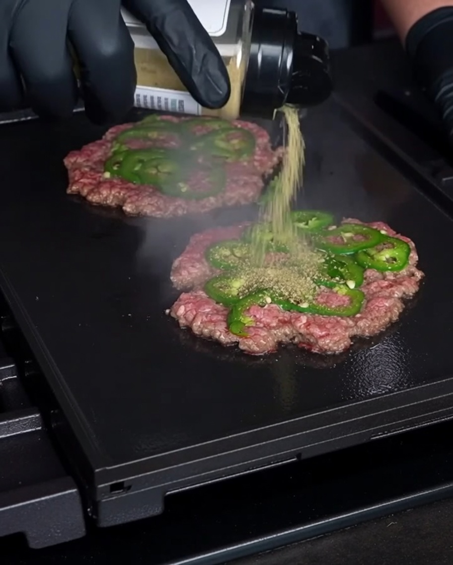 Eine behandschuhte Hand streut Gewürze auf dünne, flachgedrückte Rindfleisch-Patties, die mit frischen Jalapeño-Scheiben durchsetzt sind, auf einem heißen Grill.