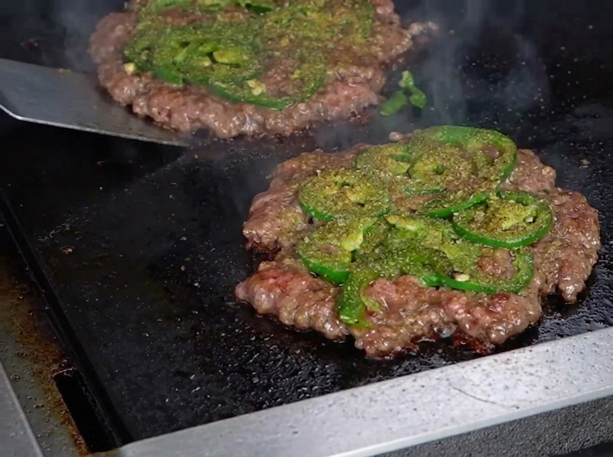 Ein Metallspatel wendet ein dünnes Smashburger-Patty und enthüllt eine perfekt gebräunte, knusprige Kruste auf der Unterseite.