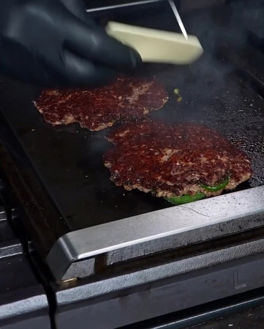 Eine behandschuhte Hand legt eine Scheibe weißen Havarti-Käse auf ein wunderschön angebratenes Smashburger-Patty auf einer heißen Grillplatte.