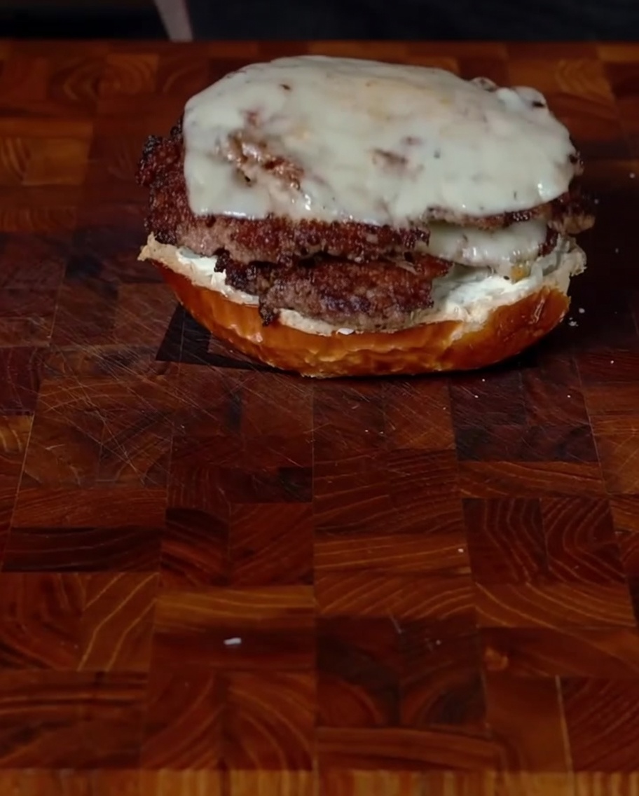 Zwei gestapelte Smashburger-Patties mit Jalapeños und geschmolzenem Käse werden vorsichtig auf ein mit Frischkäse bestrichenes unteres Bun gelegt.