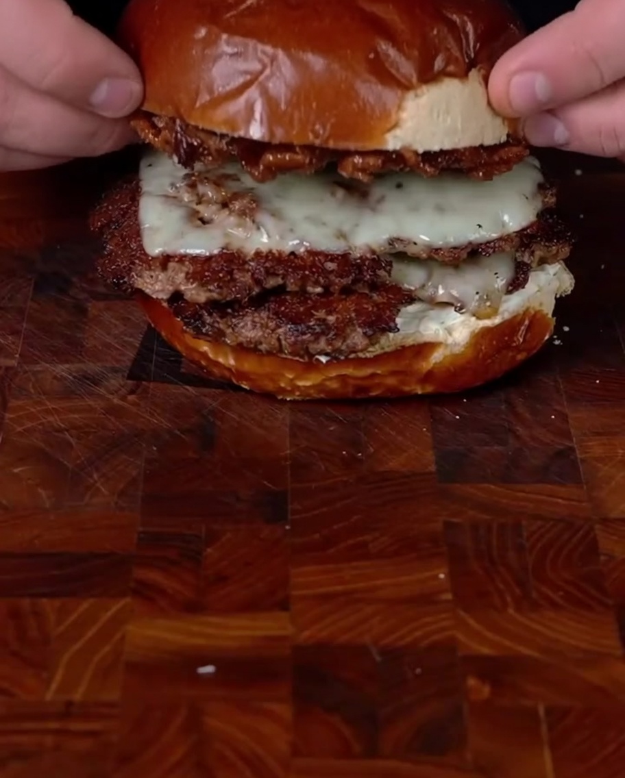 Hände setzen das obere Bun beladen mit Bacon Jam auf den gestapelten Doppel-Smashburger, um den Zusammenbau abzuschließen.