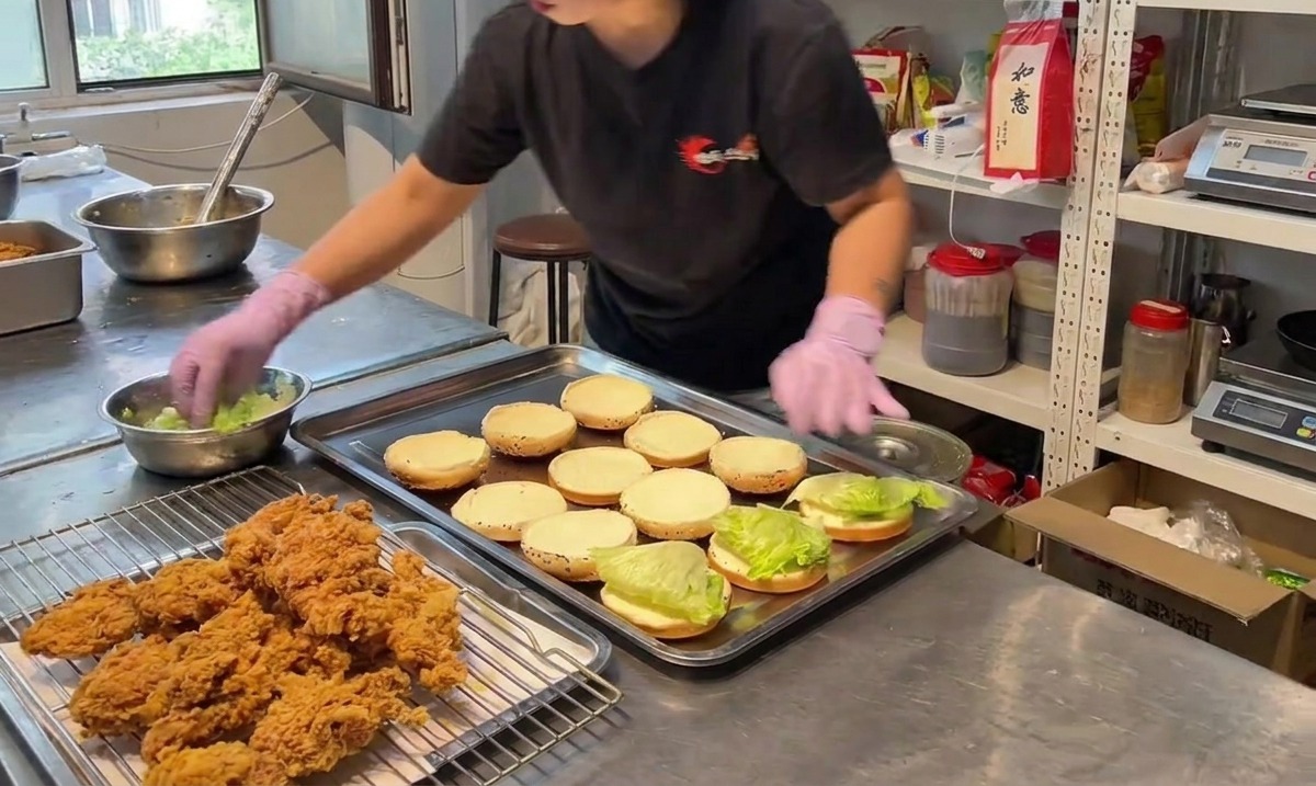 Eine Person platziert ein Stück knuspriges Hähnchen auf der unteren Burger-Brötchenhälfte mit Salat.