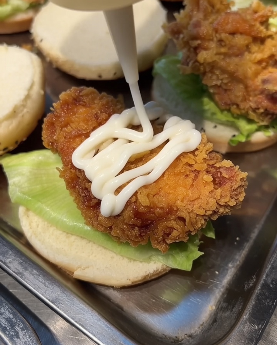 Weiße Mayonnaise wird aus einer Flasche zickzackförmig auf das Hähnchen auf dem Burger aufgetragen.