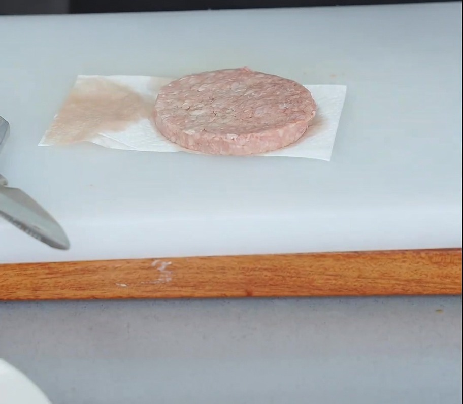 Ein rohes Rindfleisch-Patty liegt auf Küchenpapier, während ein weiteres Stück Papier darauf gedrückt wird, um Feuchtigkeit aufzusaugen.