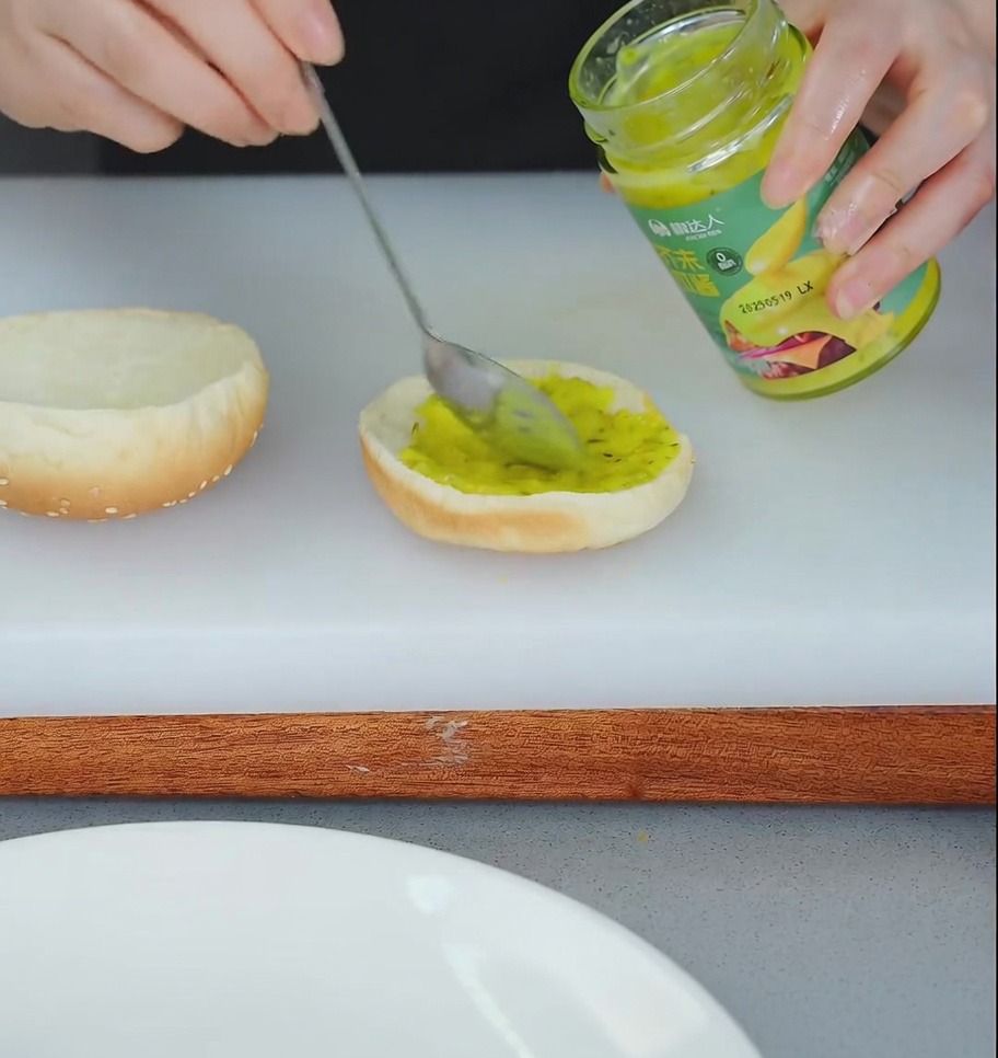 Eine Person verstreicht mit einem Löffel gelbes Relish auf der unteren Hälfte eines getoasteten Hamburgerbrötchens.