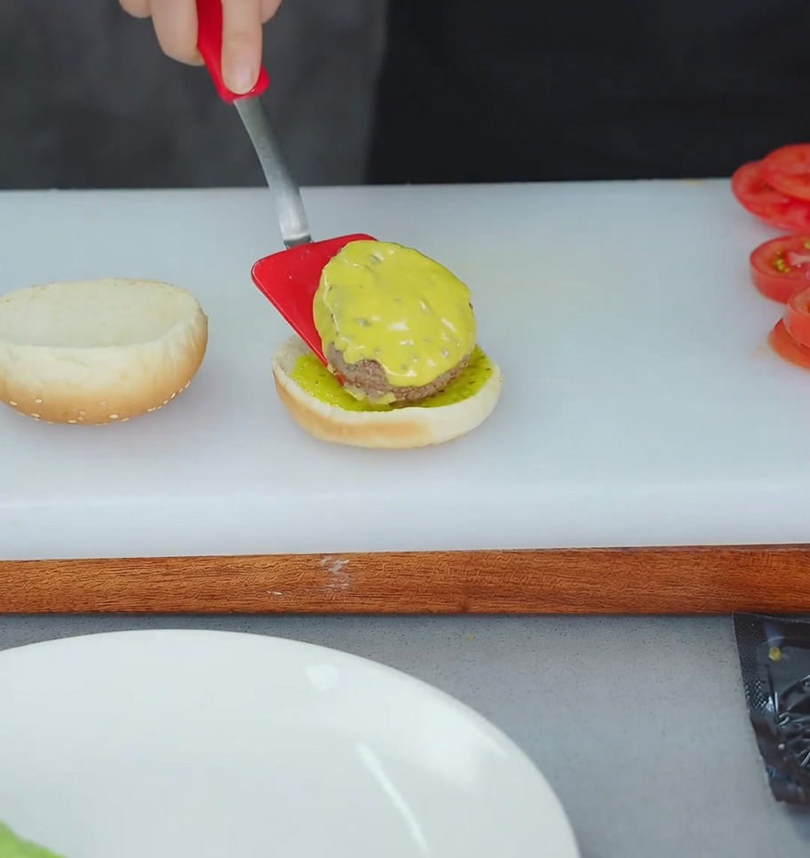 Ein roter Pfannenwender hebt ein mit Käse belegtes Rindfleisch-Patty auf ein getoastetes Burgerbrötchen mit gelbem Relish.