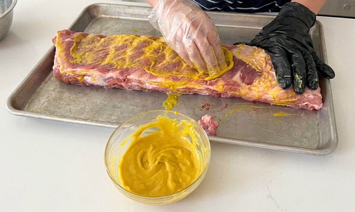 Eine Person mit schwarzen Einweghandschuhen verreibt ein Senf-Bindemittel auf rohen Schweinerippchen auf einem Backblech.