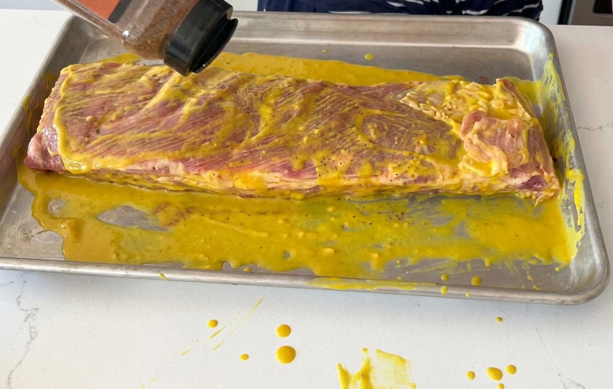 Ein BBQ-Trockenrub wird aus einem Streuer über einen mit Senf bestrichenen Rippchenstrang gegeben.