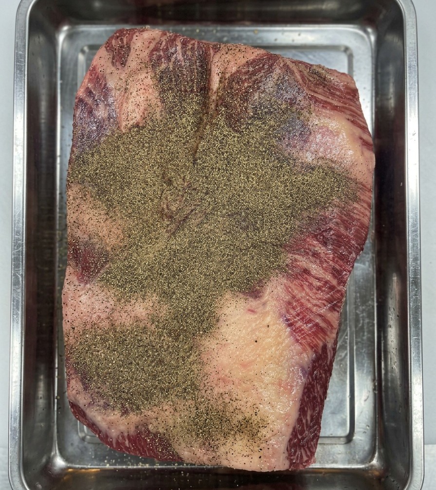 Behandschuhte Hände drücken schwarzen Pfeffer in die Oberfläche des Wagyu Briskets, um eine gleichmäßige Gewürzschicht zu erzeugen.