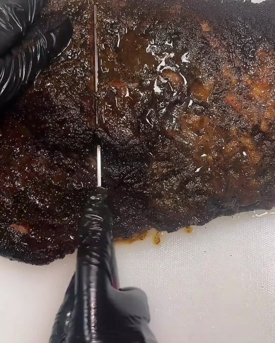 Nahaufnahme einer Person mit schwarzen Handschuhen, die mit einem langen Tranchiermesser ein dickes, dunkles geräuchertes Brisket auf einem weißen Schneidebrett anschneidet.
