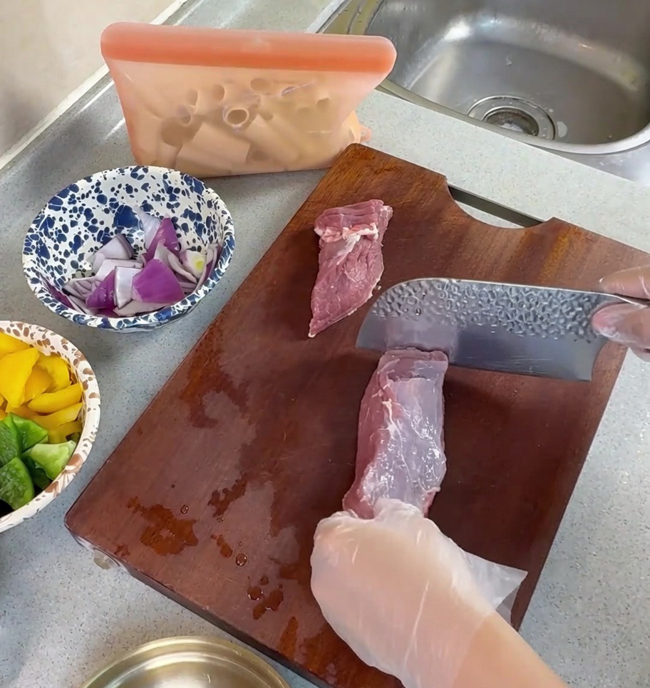 Behandschuhte Hände schneiden rohes Schweinefleisch mit einem Hackmesser auf einem Holzbrett in dünne Streifen.