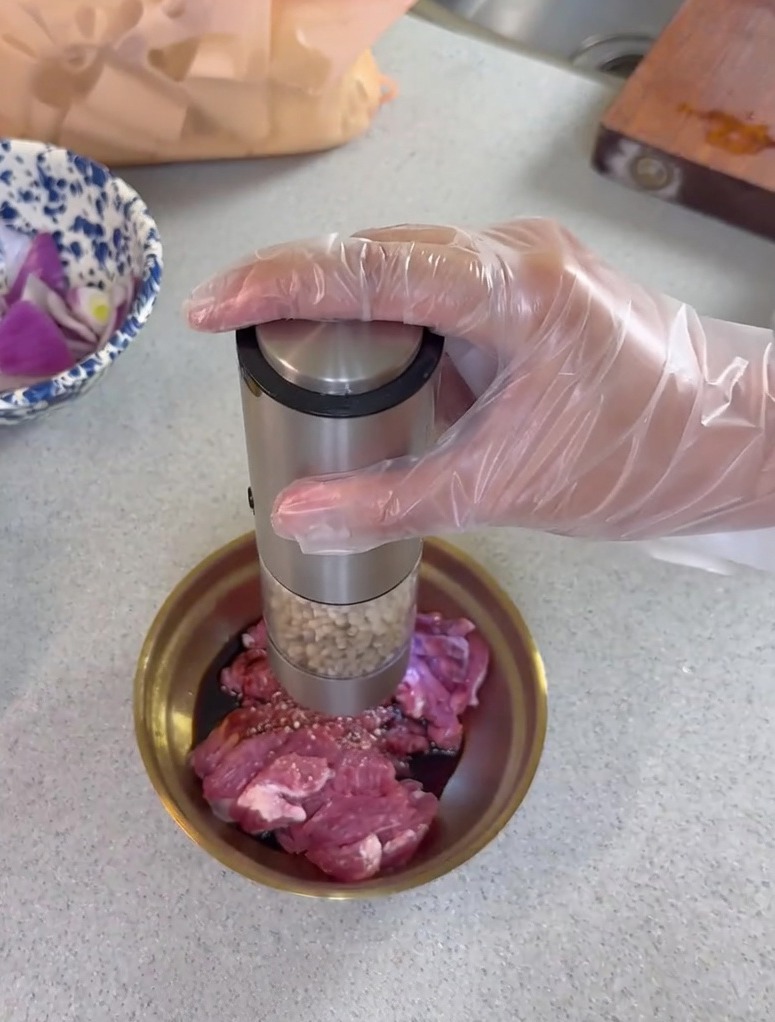 Frischer Pfeffer wird mit einer elektrischen Mühle über rohes, in einer Metallschale mariniertes Fleisch gegeben.