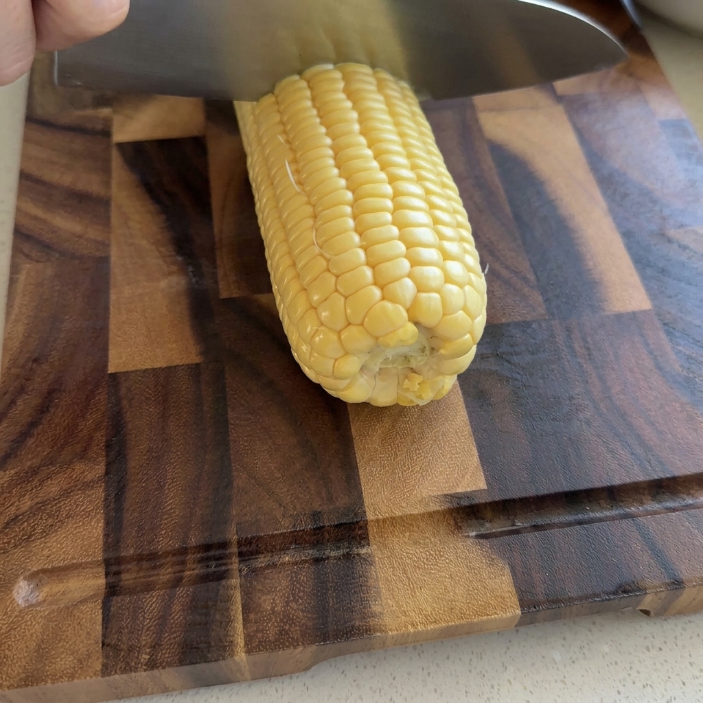 Eine Hand schneidet mit einem großen Kochmesser rohe gelbe Maiskörner auf einem Holzschneidebrett vom Kolben.