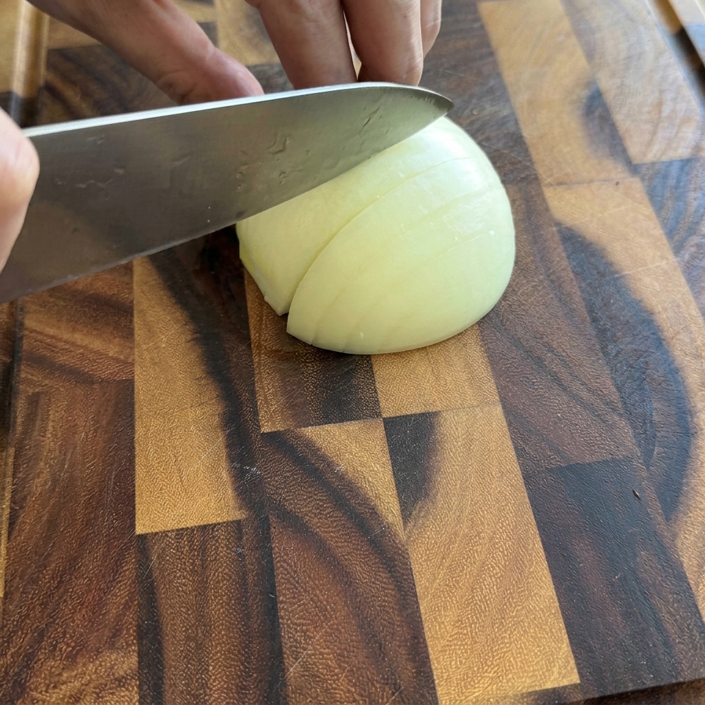Eine Person schneidet eine halbe weiße Zwiebel auf einem Holzschneidebrett in kleine Würfel.