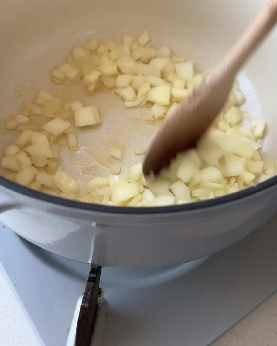 Ein Holzlöffel rührt gewürfelte weiße Zwiebeln in einem weißen Topf mit geschmolzener Butter um.