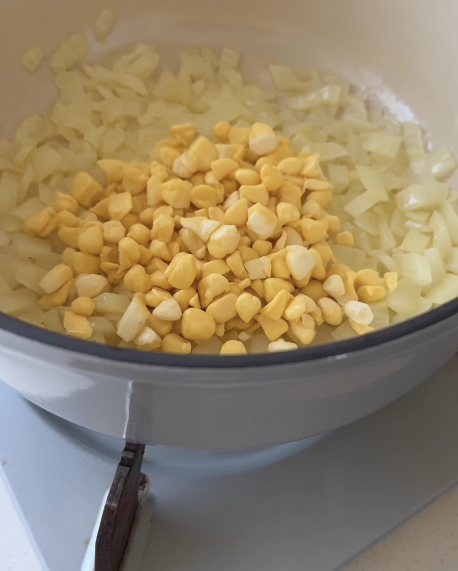 Frische gelbe Maiskörner werden in einen Topf mit glasigen Zwiebeln geschüttet.